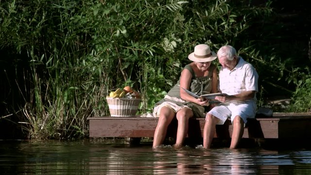 Mature couple looking at photo album. Old man and woman sitting with photo book near river.