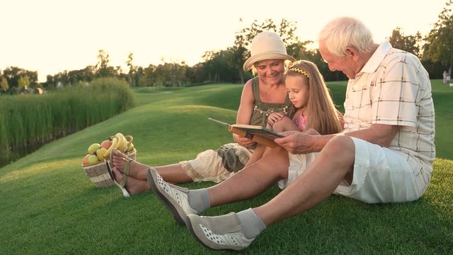 Granddaughter sitting with grandparents outdoors. Girl with her grandparents looking at photo album sitting on grass. Sweet memories in photo book.