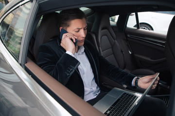 Stay always available! Young good-looking polite male sitting in the car, talking the phone and working on his wireless computer.