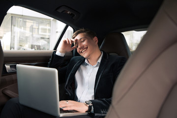 Be always cheerful and positive! Cheerful acknowledged young professional businessman sitting in the car and smiling, while working on the laptop.