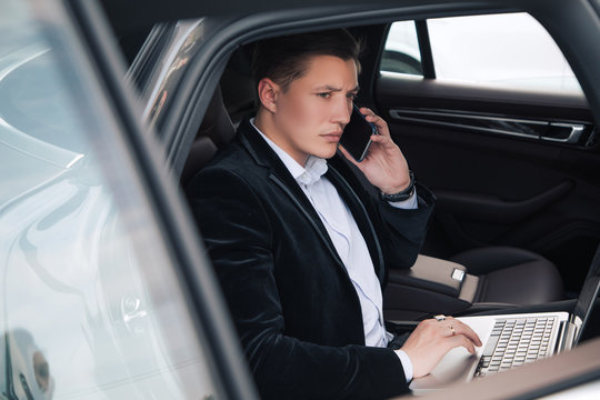 Business talks! Always online young serious handsome newcomer working on his laptop in the car and talking his smartphone.