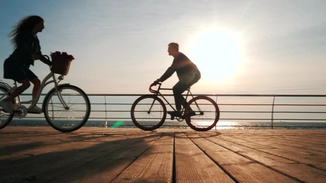 Silhouettes of young couple riding bicycles on sea embankment and driving past each other. Girl and boy on date. People and healthy lifestyle concept. Romantic scene.