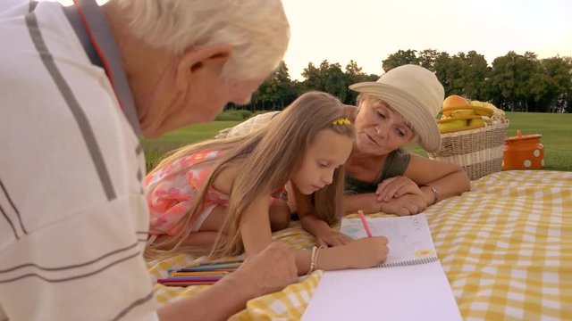 Schoolchild is drawing with colored pencils outdoors. Grandparents looking how granddaughter is drawing, summer nature background. Support of relatives is priceless for skills development of kids.