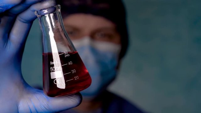 Portrait of a doctor in blue workwear, holding a flask with analyzes, blue rubber gloves, blue hat, blue diffuser, blue background. Concept: doctor, herurg, chemist, trainee.