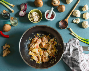 Chicken pieces with spices in wok frying pan on kitchen table with vegetables ingredients and wooden spoon, top view. Cooking preparation