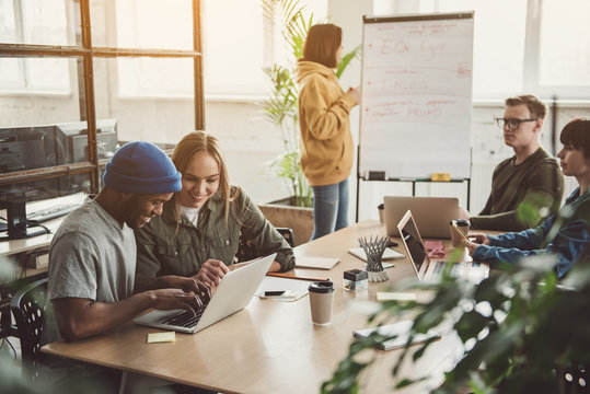 Beaming girl and cheerful male looking at laptop while having job with partners in office. Female describing information while locating near board. Start up team concept