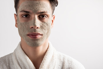 Fototapeta premium Skin care concept. Portrait of serious young man with dry clay mask on face is standing against light wall and looking at camera thoughtfully. Spa and beauty treatment. Copy space in the right side