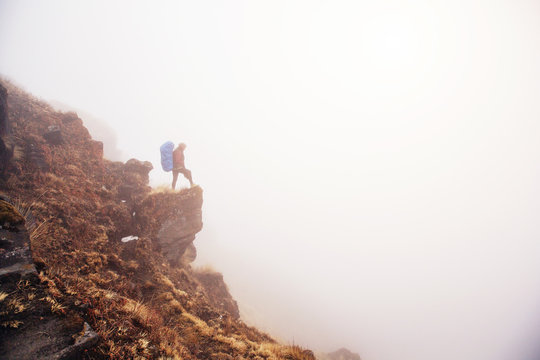 Female traveller with big backpack staying on the top of mountain cliff in white fog above clouds and valley, enjoy view