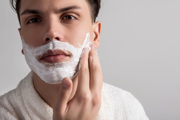 Close up of face of young attractive man in bathrobe is applying shaving foam on cheeks while having morning skincare. Copy space in the right side. Daily hygiene concept