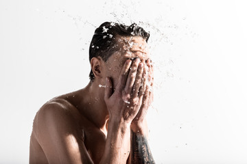 Side view of young naked muscular guy is standing against light background and spraying clean water...