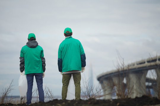 Backs of two guys in green uniform standing in front of littered territory against grey sky