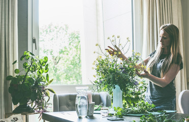 Beautiful woman with long blond hair arranging wild flowers bunch in vase on table in living room...