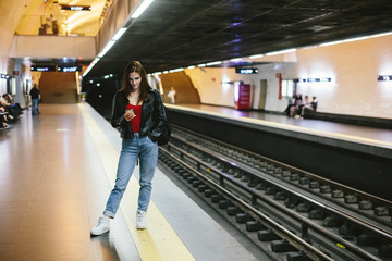 Young casual female waiting for train in metro station, using mobile phone