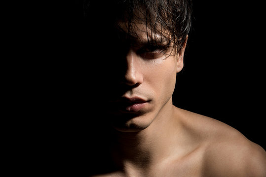 Close up portrait of young mysterious guy with half face in shadow. He is standing on dark background and expressing thoughtfulness. Masculine beauty concept