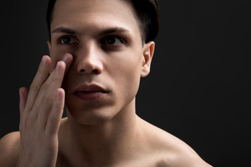Avoid wrinkles around eyes concept. Close up portrait of pleasant shirtless guy is carefully putting moisturizing cream on his face by fingers. Copy space in the right side