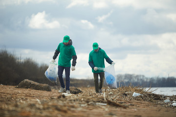 Two men with big sacks walking along river bank, picking up litter and putting it in those sacks
