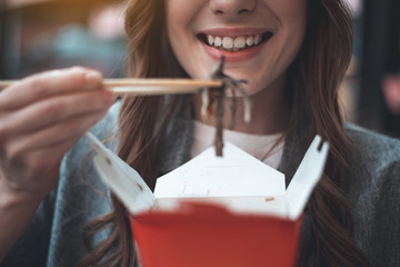 Focus on lady grin enjoying asian meals. She is taking dish of take-away box using chopsticks. Close up