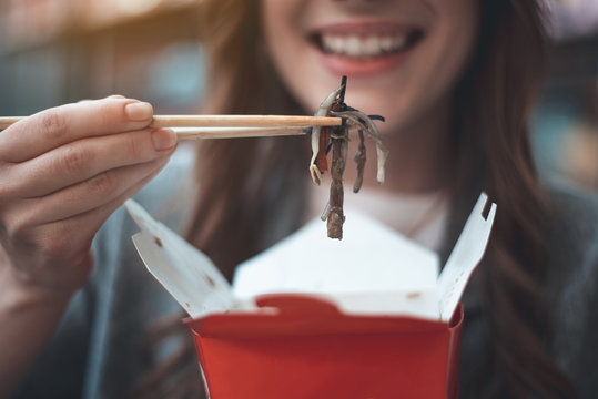 Focus on woman hand holding oriental meal with chopsticks. She is taking dish out of take-away box. Girl is smiling. Close up