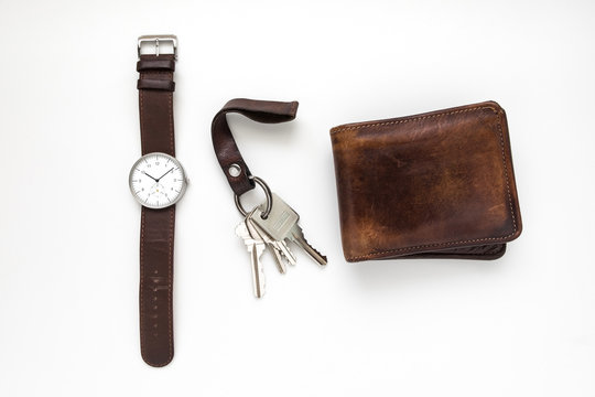 Top view of everyday carry objects made by brown leather wrist watch, key chain, wallet on white background