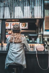 Lady is sitting at counter in cafeteria with back to camera. She is wearing trendy hat and putting purse on the table
