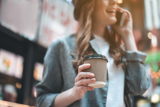 Low angle focus on woman hand holding cup of coffee. Cheerful girl is enjoying phone conversation