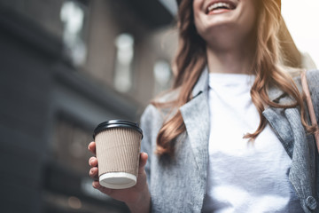 Low angle close up of female hand holding hot drink outside. Lady is standing and smiling somewhere outdoors