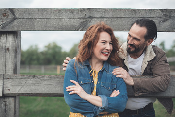 Cheerful married couple is having rest in countryside. They are standing near wooden barrier and...