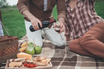 Cercles muraux Pique-nique Gros bras de l& 39 homme versant du vin rouge de la bouteille dans le verre. La femme est assise près de son mari sur la couverture et se détend. Pique-nique romantique sur le concept de terrain en herbe
