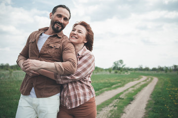 You are my love. Waist up portrait of joyful red-haired woman is cuddling her husband with fondness. They are standing on meadow alley and laughing. Copy space 
