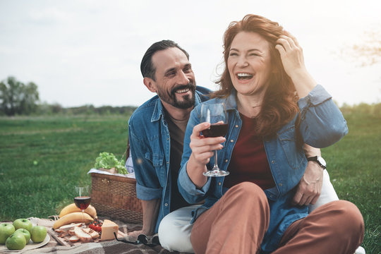 Wonderful day for being together. Portrait of excited loving couple having romantic picnic on the meadow. They are embracing and laughing - Powered by Adobe