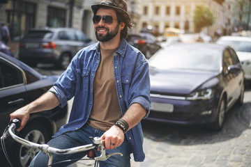 Fototapeta na wymiar Life in movement. Portrait of cheerful man in headwear and sunglasses riding bike along the road. Cars on background. Copy space in right side