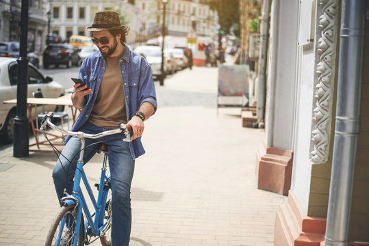 Full length portrait of good looking male person coming to stop during cycling. He is looking at mobile phone in his hand with smile. Copy space in right side
