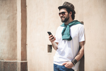 Fototapeta premium Cheerful man with modish appearance standing against the wall outside and staring with joy. Smartphone is in his hand. Copy space in left side