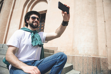 Low angle of young trendy male person making selfie on mobile phone while resting out of doors. He is smiling. Copy space in right side