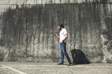 I can not imagine life without smartphone. Side view profile of bearded young man walking down the street and looking at his mobile phone. Concrete wall on background. Copy space in left side