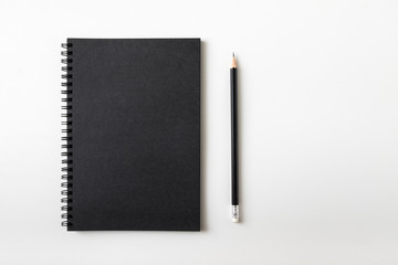Top view of closed black cover notebook with pencil on white desk background