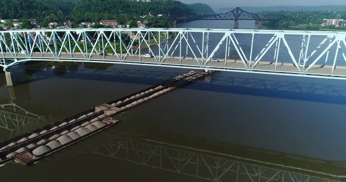 A morning aerial establishing shot of a coal barge traveling under a bridge on the Ohio River. Reverse shot, Pittsburgh suburbs.  	