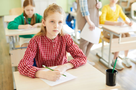Serious schoolgirl sitting by individual desk and looking at blackboard while carrying out test