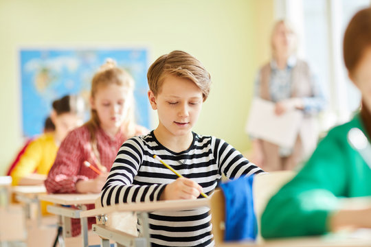 Schoolboy and his classmates sitting by individual desks and carrying out exam assignments at lesson