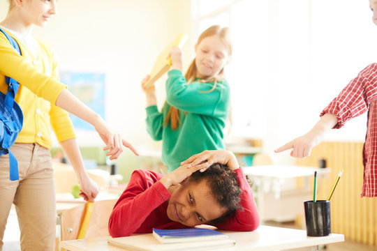 Two schoolgirls pointing at one of their classmates while girl with book going to hit him