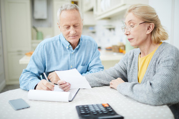 Senior couple sitting by table in the kitchen and planning family budget for various expenses