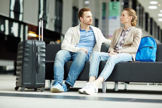Young modern couple sitting in airport lounge and having talk while waiting for airplane