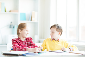 Two friendly little schoolchildren sitting by desk in classroom, thinking of ideas and discussing them