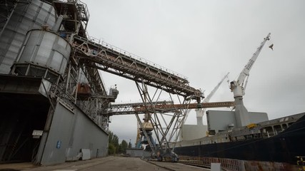 Timelapse of grain terminal at seaport on cloudy day. Cereals bulk transshipment to vessel loading grain crops on bulk ship from large elevators at the berth. Transportation of agricultural products.