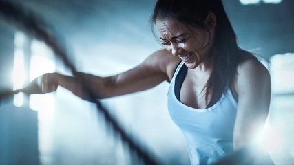 Athletic Female in a Gym Exercises with Battle Ropes During Her Cross Fitness Workout/ High-Intensity Interval Training. She's Muscular and Sweaty, Gym is in Industrial Building.