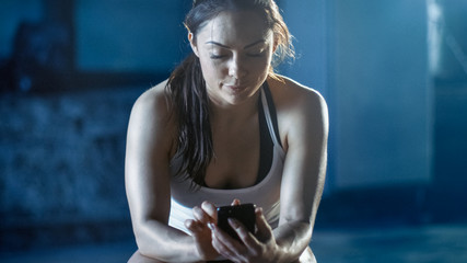 Fototapeta na wymiar Beautiful Athletic Brunette Uses Smartphone while Resting on the Bench after Her Intensive Cross Fitness Bodybuilding Gym Training.