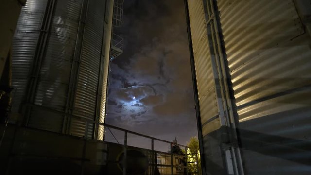 Timelapse of grain terminal at night. Moon and clouds in the sky. Metal tanks of elevator. Grain drying complex construction. Commercial seed silos at seaport. Steel storage for agricultural harvest.