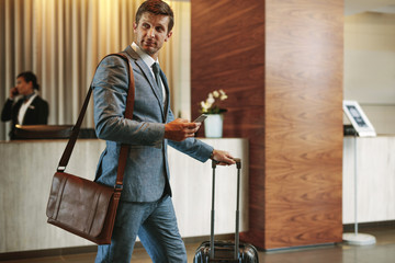 Businessman in hotel hallway with phone and baggage - obrazy, fototapety, plakaty