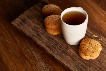 White porcelain mug of tea and sweet cookies on piece of wood over wooden background, top view, selective focus