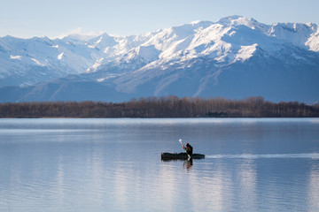 Canoe on a lake with beautiful snowy mountains on the background. Winter panorama with male paddler padding on the blue cold water. A man canoeing alone. Kayak on the river in a sunny day.